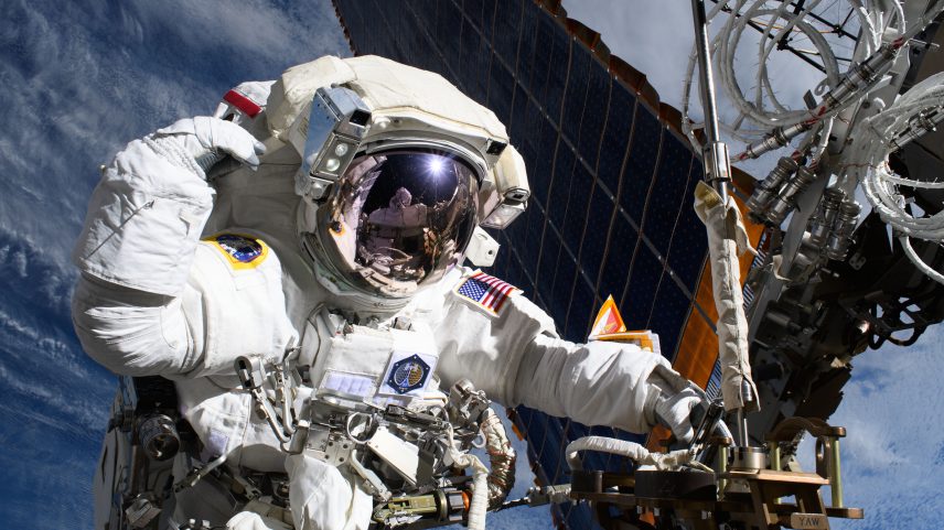 An astronaut outside of the International Space Station has one hand on a truss near a solar panel. Her other hand is by her head. Reflected in her helmet is astronaut Nichole Ayers, also in a white spacesuit, taking the photo. Earth's blue water and white clouds can be seen in the background.