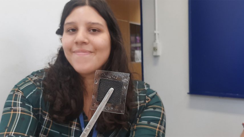 Protótipo da bateria flexível, na qual a parte transparente é uma membrana de troca de prótons que substitui o meio líquido de uma bateria convencional e a parte escura são as nanopartículas de chumbo que constituem os eletrodos, positivo de um lado e negativo do outro. Ao fundo, a doutoranda Victória Amatheus Maia – Foto: Ivan Conterno