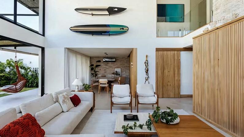 LIVING | As duas pranchas de surfe do acervo dos moradores foram fixadas no alto da parede e ganharam destaque com o pé-direito duplo. Esquadrias de PVC preto ebanizado, da Esquadrias Macaé. Tapete da Galeria Hathi. Marcenaria de lâmina de carvalho natural, da Mag Marcenaria. Sofá Blend, da Way Design. Poltrona de madeira da Empório Casa. Mesas de centro da Arquivo Contemporâneo. Almofadas Codex Home. Piso de porcelanato Super Quadra Cru, da Portobello. Revestimento da parede de tijolo Hole Stone, da Palimanan. Obras de arte e adornos da Calezani Design