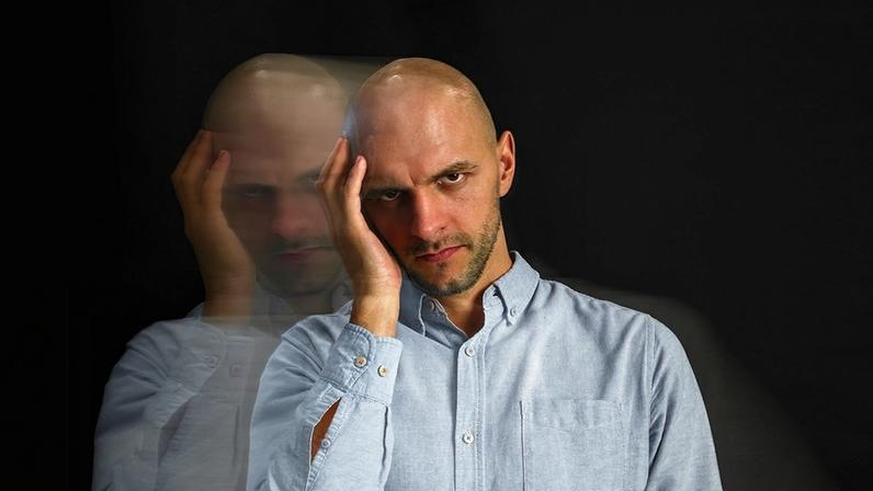 foto em preto e branco, com fundo preto, mostra um homem com uma das mãos no rosto e, ao seu lado, aparece a mesma imagem borrada