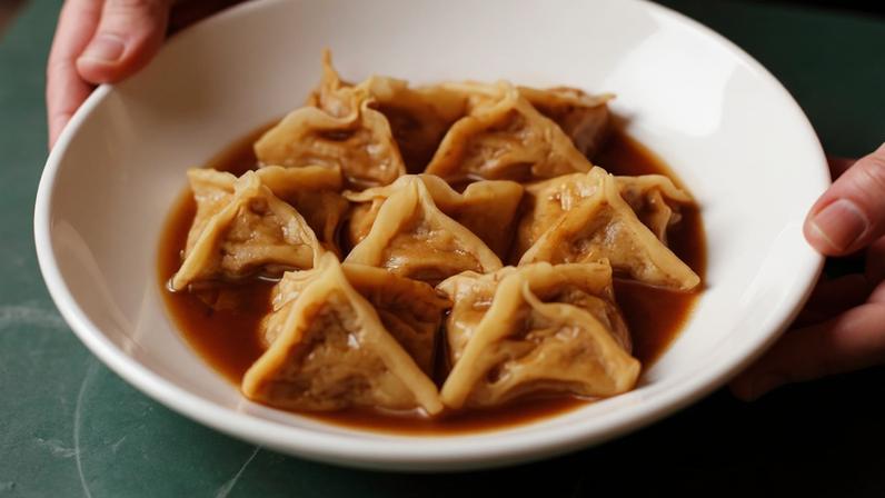 Um prato redondo contendo dumplings em formato triangular, mergulhados em um molho escuro. Os dumplings estão dispostos em uma única camada, com um molho ao redor. As mãos de uma pessoa seguram o prato.