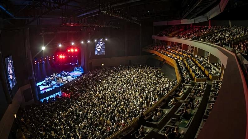 A imagem mostra um grande auditório lotado durante um show. O palco está iluminado com luzes vermelhas e brancas, e há uma banda tocando. O público é visível em várias seções, com muitas pessoas em pé e aplaudindo. As arquibancadas estão cheias, e há telões visíveis ao lado do palco.