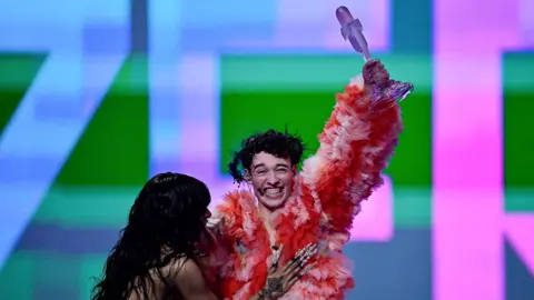 Getty Loreen and Nemo embrace as Nemo holds the Eurovision Song Contest trophy aloft. Loreen has long black curly hair and long silver nails, Nemo is wearing a pink and red ruffled coat