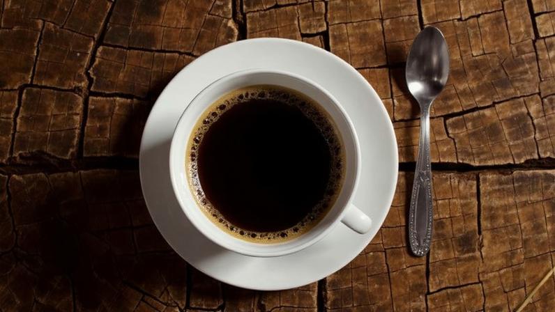 A cup of coffee with beans and a teaspoon on a stump tabletop. Image by Anja (cocoparisiene) from Pixabay (Pixabay Content License).