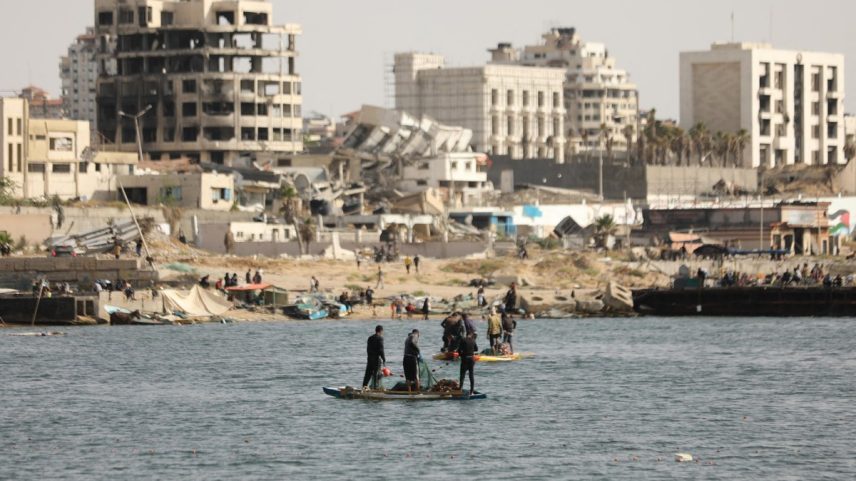 Pescadores palestinos tentam pescar com barcos a remo e varas de pescar na costa da Cidade de Gaza, enquanto prédios danificados e destruídos são vistos ao fundo (Foto: DAWOUD ABO ALKAS / AFP).