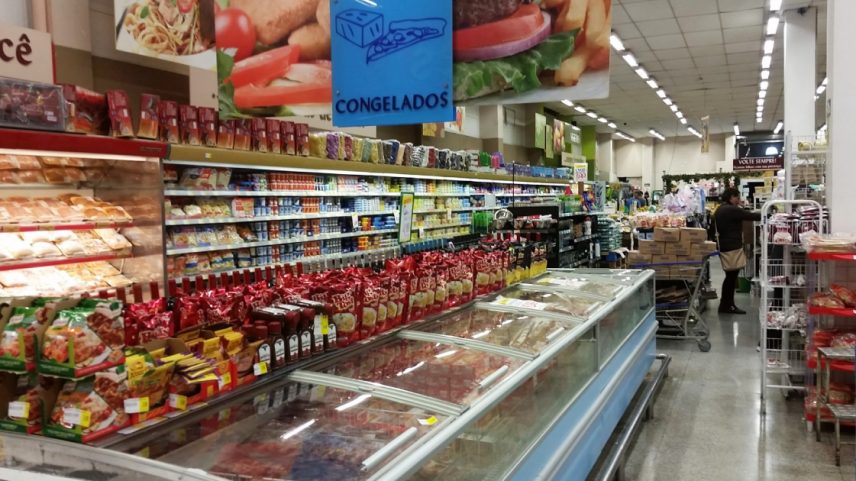 Corredor de supermercado com cartazes mostrando alimentos, prateleiras repletas de alimentos processados e embalados e uma geladeira horizontal com produtos congelados (Foto: USP Imagens)