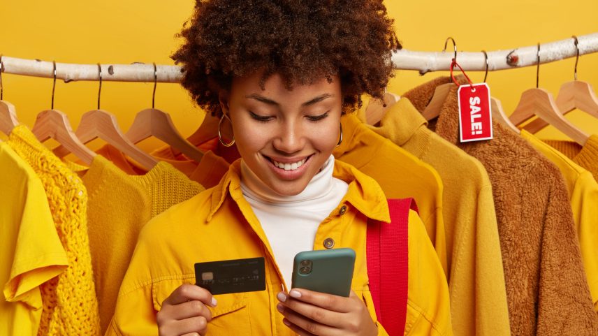 Mulher de pele escura usa celular moderno e cartão de crédito, faz compras online, realiza pedido pela internet, insere informações da conta bancária, está em frente a araras de roupas. Conceito de grande liquidação (Foto: Freepik)