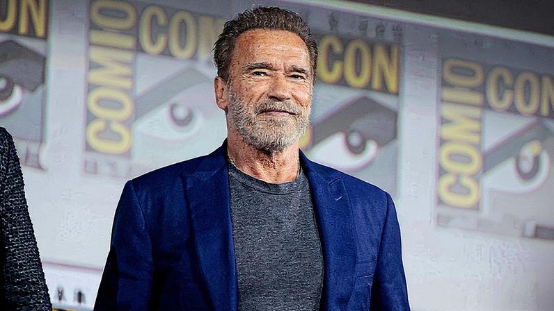 Homem em pé, vestido com um terno, fala ao microfone em um palco, com um gráfico azul em exibição ao fundo (Foto: Reprodução)