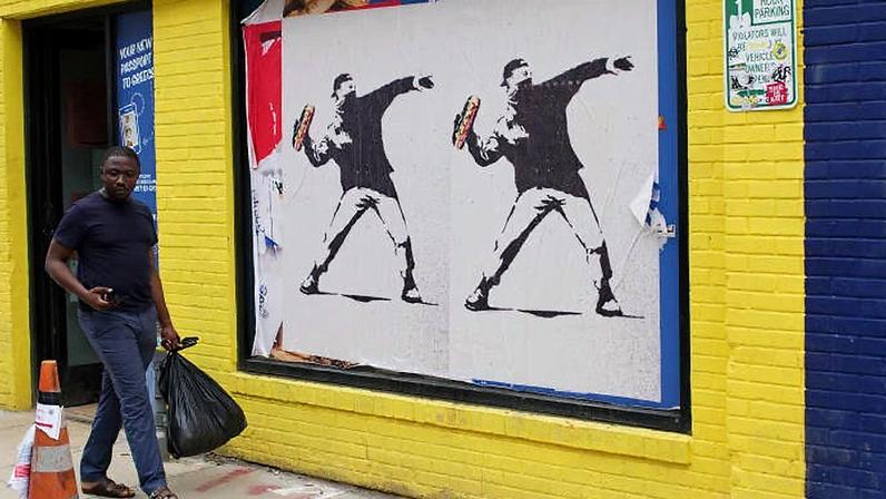 Uma pessoa passa por pôsteres no estilo Banksy de um manifestante jogando um sanduíche em 21 de agosto de 2025 em Washington, DC. (Foto: Kevin Dietsch | Getty Images)