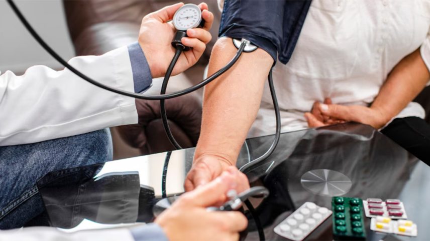 Braço sobre uma mesa usando um aparelho de medir a pressão. Os braços de uma pessoa de jaleco manipulam o aparelho. Alguns comprimidos estão sobre a mesa. (Foto: USP Imagens)
