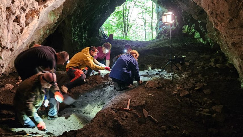 Seis pessoas agachadas trabalhando com ferramentas na escavação do solo no interior de uma caverna (Foto: USP Imagens)