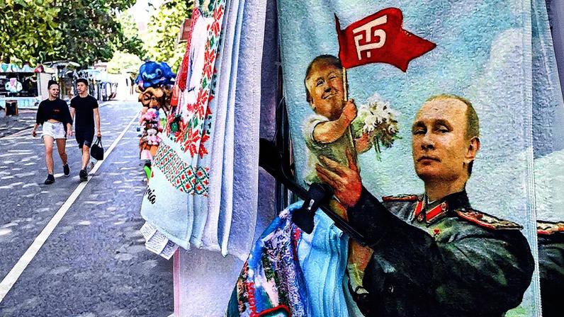 Turistas passam por barraca vendendo toalha de praia retratando o líder da Rússia, Vladimir Putin, segurando um bebê Donald Trump, presidente dos EUA, no resort de Albena, no Mar Negro (Foto: Nikolay DOYCHINOV / AFP).