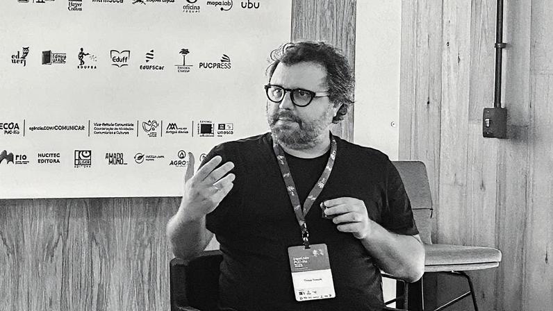 Homem de terno fala ao microfone durante apresentação em um palco com gráfico ao fundo (Foto: Reprodução)