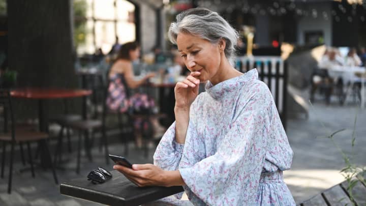 Uma mulher idosa olhando para o celular em um café na calçada. (Foto: Yuliya Taba | E+ | Getty Images)