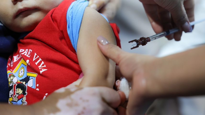 Criança olha para uma enfermeira que se prepara para aplicar uma vacina (Foto: USP Imagens)