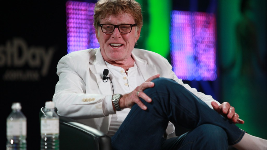 Homem de terno em pé em um palco, falando ao microfone com um grande telão ao fundo exibindo um gráfico azul em ascensão (Foto: Reprodução)