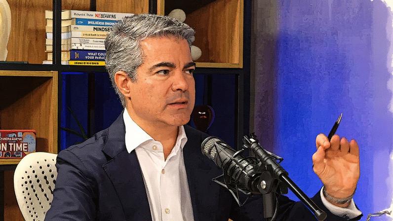 Homem em terno fala ao microfone durante apresentação em um palco com gráfico ao fundo (Foto: Reprodução)