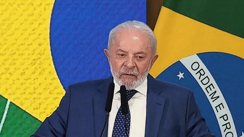 Homem em terno fala ao microfone durante apresentação em um palco com gráfico ao fundo (Foto: Reprodução)