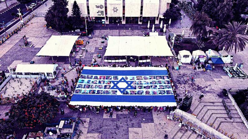 Protesters in Habima Square, also known as Hostages Square, a public plaza located in front of the Tel Aviv Museum of Art, hold up an Israeli flag featuring photos of the hostages held in Gaza, demanding their return. Photo: Yair Palti/Anadolu via Getty Images. © 2025 Anadolu