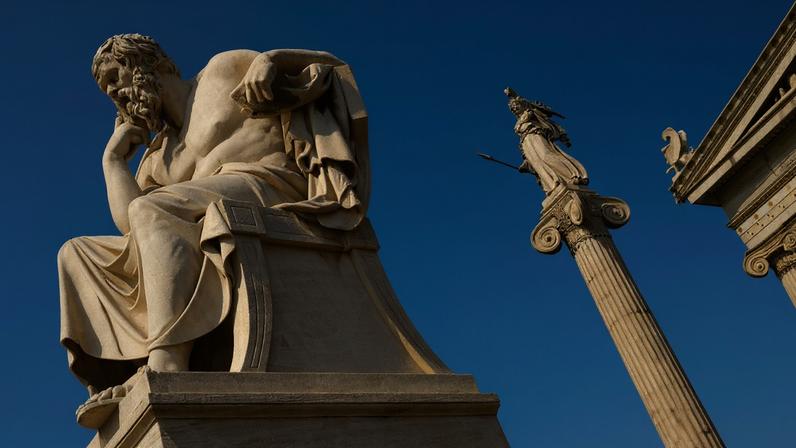 Marble statues of Socrates seated in thought and a standing Athena atop a tall column, set against a bright blue sky.