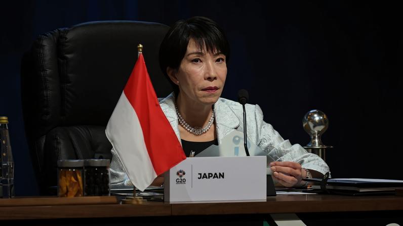 Takaichi sits at a desk with a microphone behind a placard that says "Japan." She wears a white business jacket and is holding a plastic folder.