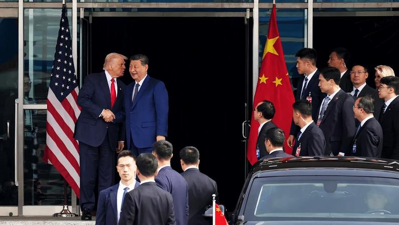 Trump and Xi stand in the exterior doorway of a building, shaking hands as Trump leans over to whisper something in Xi's ear. Aides and other officials stand nearby watching. All wear formal business suits.