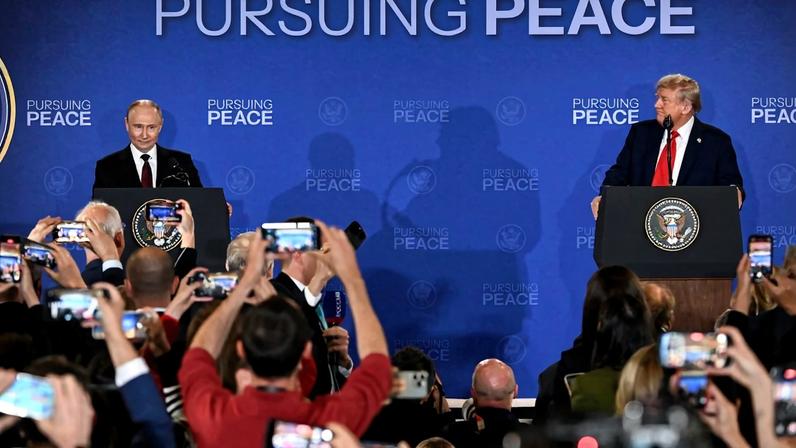 Putin and Trump stand at podiums on a low stage, about ten feet apart. Putin smiles while Trump is turned to the side to watch him. Journalists are visible in the crowd in front of the stage, holding phones, video cameras, and microphones as they record.
