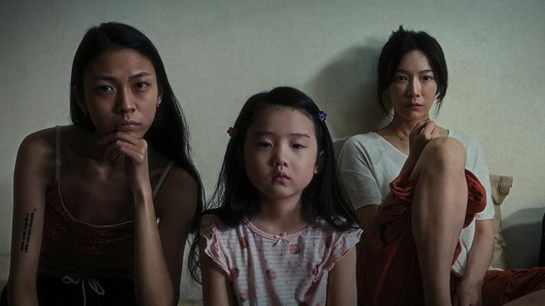 A young girl is flanked by two women, all with serious faces.
