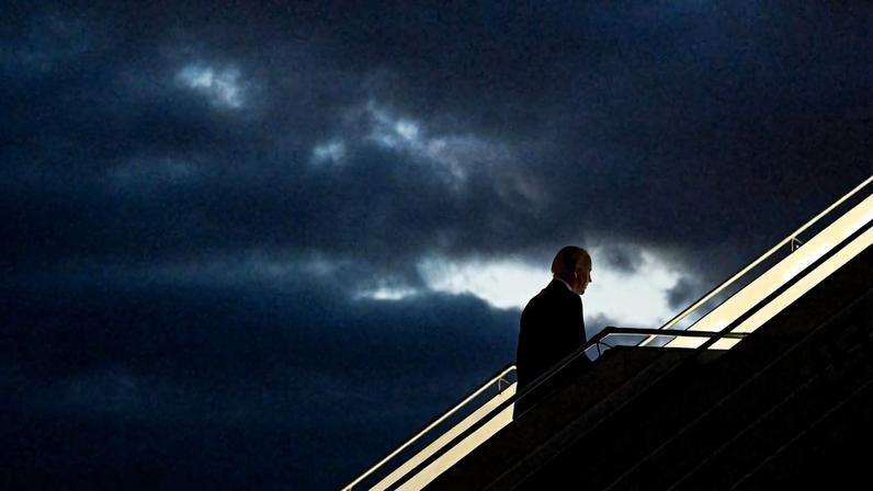 Joe Biden, in silhouette, walks up stairs to board a plane, with ominous dark clouds in the background.