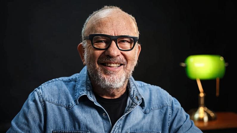 Homem idoso com barba branca e óculos de armação preta sorri para a câmera. Ele veste uma camisa jeans azul clara sobre camiseta preta, com fundo escuro e luminária verde desfocada ao fundo.