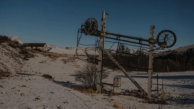 An old ski lift stands on a slope on which grass can be seen poking through a light covering of snow.