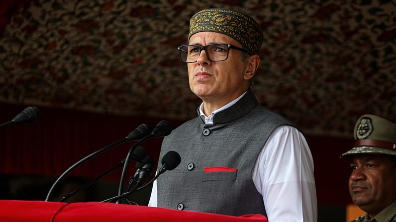 Omar Abdullah wears black-rimmed glasses, a white long sleeve shirt under a grey vest, and a grey and gold skullcap. He stands behind a red podium and gazes out with a serious expression toward his out-of-frame audience.