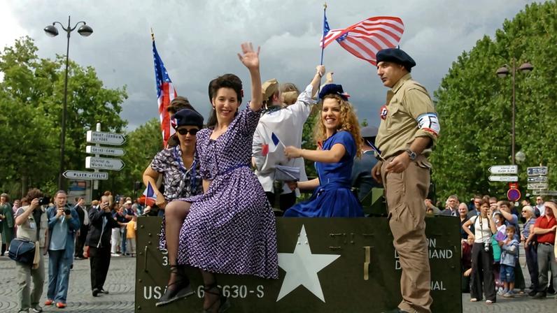 Two women wearing 1940s-style dresses and hair and makeup perch in the back of a military truck, waving toward the camera. Other people are dressed in berets and military uniforms or khaki outfits to represent the French resistance. The truck drives down the middle of a road away from the camera between spectators on either side, a cloudy sky looming overhead.