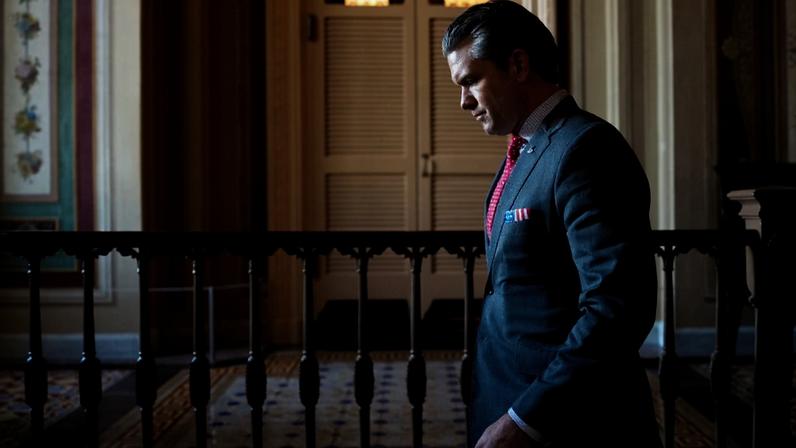 Hegseth is seen in profile as he walks through a hallway on Capitol Hill. He wears a dark suit and red tie, and his face his partly in shadow.