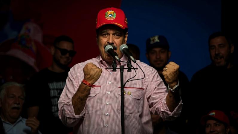 Maduro speaks into a microphone as he stands with his fists raised while wearing a red baseball cap and a red-and-white striped button-down shirt. People onstage behind him smile toward the camera.