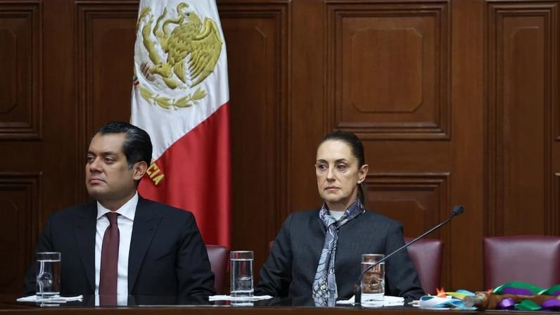 Sheinbaum sits next to Gutiérrez at a desk in front of a wood-paneled wall. Between them and the wall is a Mexican flag. Both wear business formal attire and serious expressions on their faces.