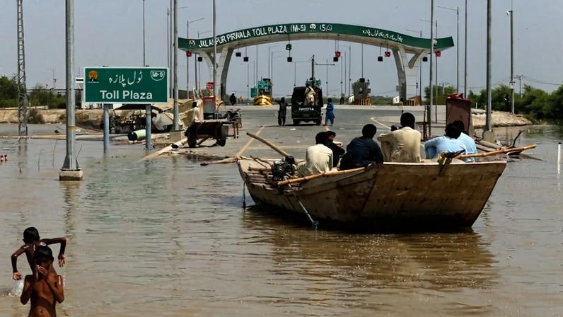 People sit on a boat on a flooded road near a toll plaza.