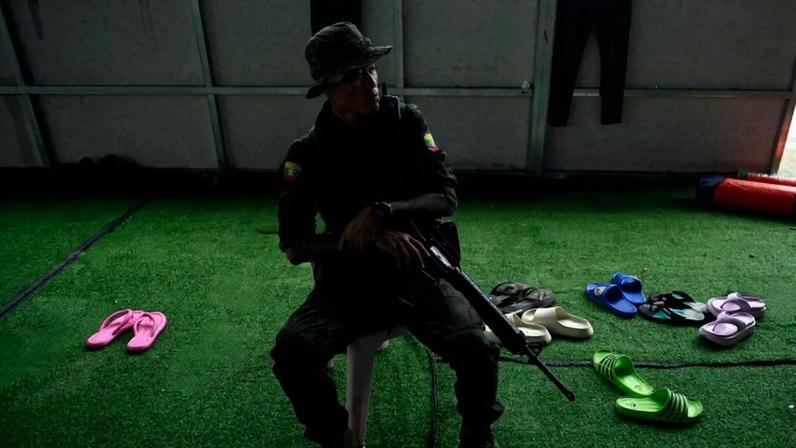 A guard with a gun sits in a chair in the shadows. Behind him are dozens of sandals.