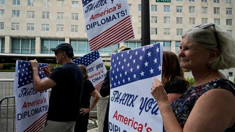 A smiling woman holding a sign that reads "Thank you America's diplomats" stands in the foreground of a group of supporters outside of the U.S. State Department.