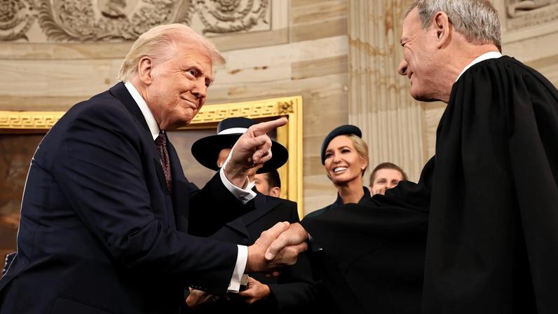Trump smirks as he leans forward and shakes Justice Roberts's hand. With his free left hand, Trump jabs one finger and points at Roberts. Other people in formal attire smile and look on.