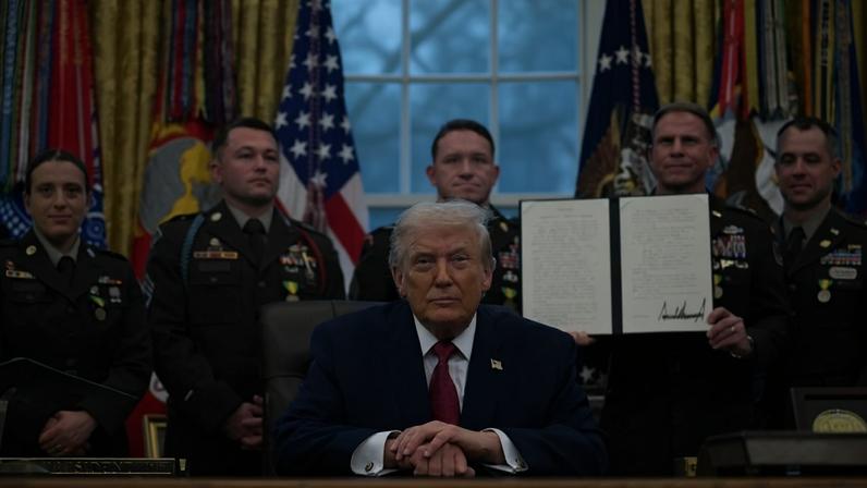 Trump sits at the Resolute Desk in the Oval Office with his hands clasped in front of him. He wears a dark suit and red tie. Military officers in dress uniform stand behind him, with one of them holding an open folder showing a document with Trump's signature at the bottom.