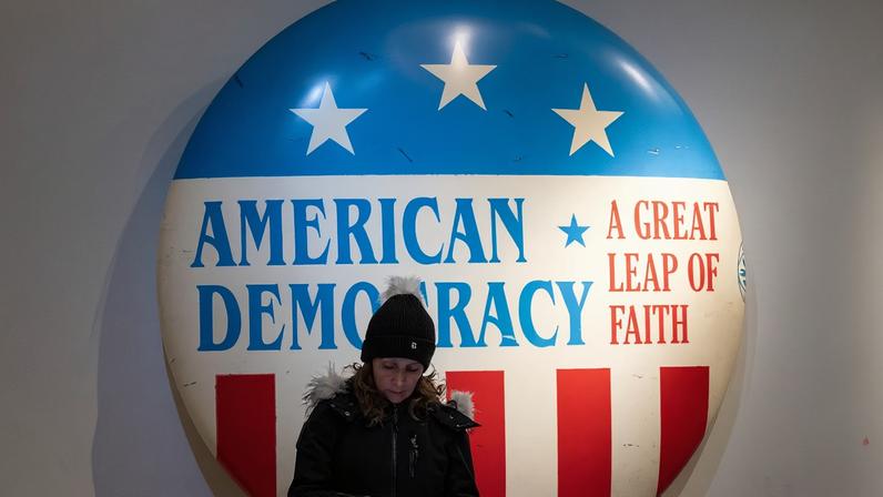 A woman stands in front of a large wall-mounted red-white-and-blue button that reads "American Democracy: A Great Leap of Faith."