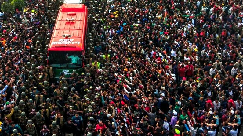 An overhead shot shows a crowd of hundreds of people cheering and reaching their arms toward a red bus parked in the middle of the crowd. Rahman stands inside at the front of the bus, visible through the windshield as he lifts his arms and waves. Security personnel in helmets and vests cluster close around the car.