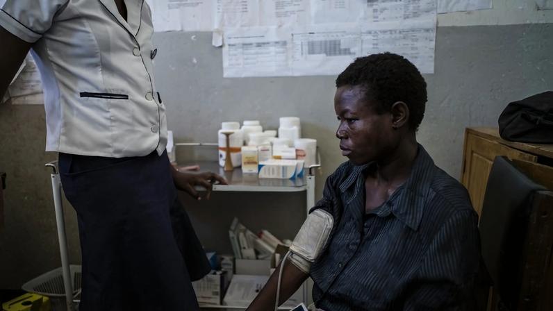 A woman sits in a clinic exam room with a blood pressure cuff on her arm, looking to the side with a serious expression. A nurse in a white uniform shirt stands nearby. Medical supplies and medication bottles are arranged on a small shelf against the wall, which is covered with charts and papers.