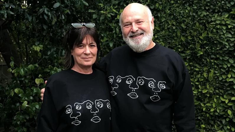 Rob Reiner and his wife Michele standing outside wearing matching black sweatshirts with line art faces.
