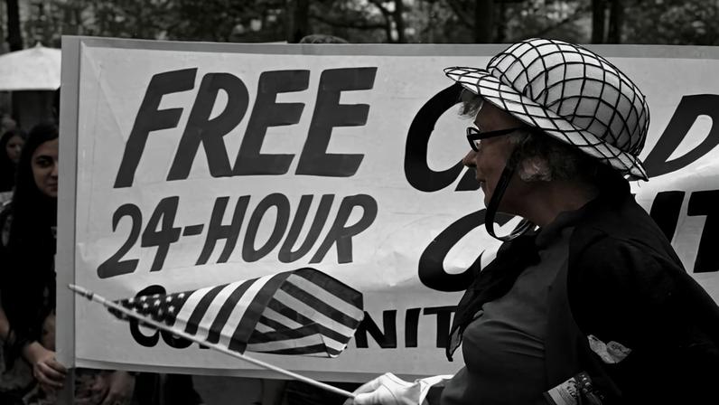 In this black-and-white photo from the 1970s, a woman wearing a pith helmet with a net over it waves an American flag as she stands in front of a banner that reads “Free 24-Hour Child Care Centers Community Controlled.”