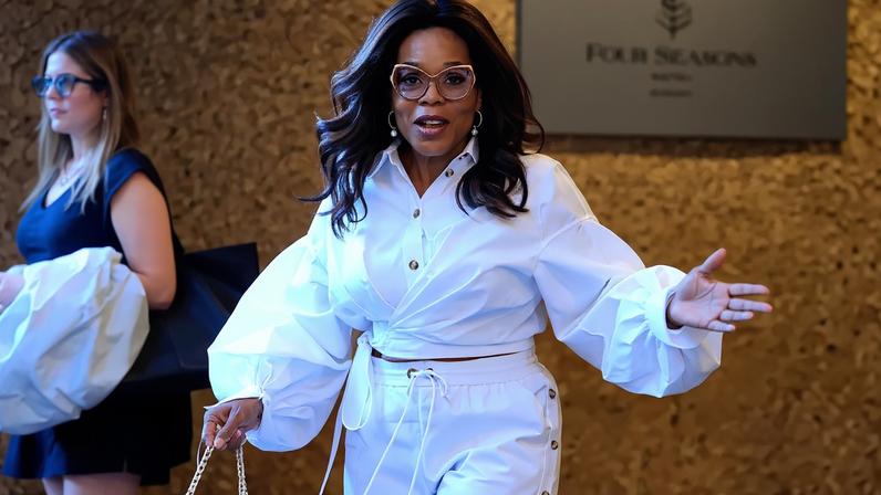 Oprah Winfrey in an all-white ensemble, including a cropped top and wide-leg pants with snap-button detail, sunglasses, hoop earrings, and a pink handbag, in Paddington.