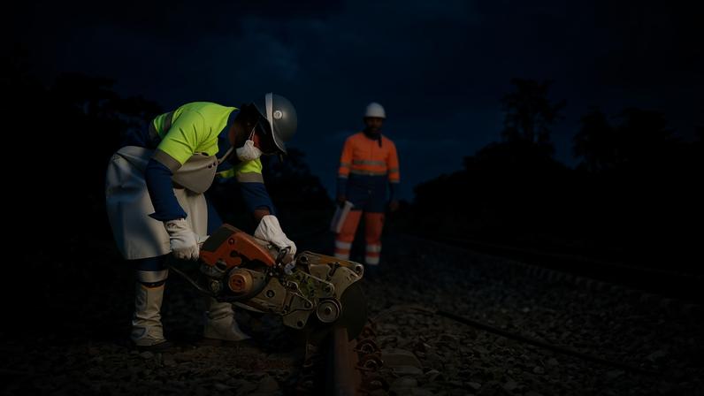 A man in the foreground bends over construction equipment he is using to renovate a section of railroad while his coworker looks on in the background in a isolated and forested area of Gabon.