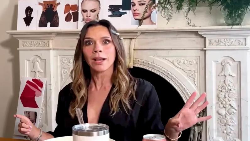 Victoria Beckham sitting at a table with an assortment of makeup inspiration images on a board behind her.