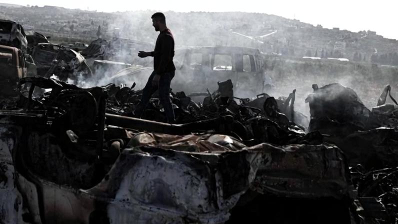 A man walks through a wide scene of the charred and smoking remains of burnt cars.